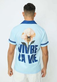 Chemise bleu clair avec un col bleu, présentant un grand motif de fleur et un texte en gras "VIVRE LA VIE" au dos, manches courtes, texture lisse.