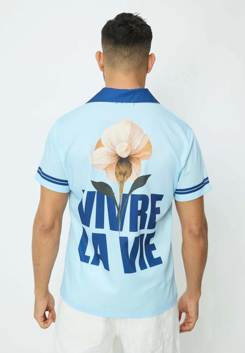 Chemise bleu clair avec un col bleu, présentant un grand motif de fleur et un texte en gras "VIVRE LA VIE" au dos, manches courtes, texture lisse.