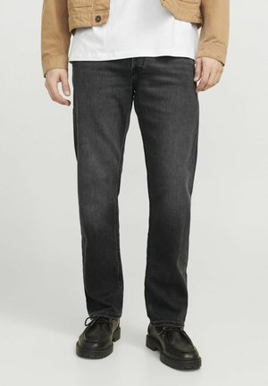 Man die zwarte jeans met rechte pijpen, zwarte leren schoenen, een wit T-shirt en een lichtbruine jas draagt, staand voor een neutrale achtergrond.