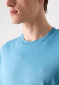 Blaue Baumwoll-T-Shirt mit Rundhalsausschnitt, ausgestattet mit einem dezenten eingeprägten Logo auf der linken Brust und einer glatten, weichen Textur.