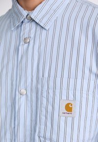 Chemise boutonnee bleu clair avec des rayures verticales. Présente une poche sur la poitrine et une étiquette logo Carhartt. Fabriquée en tissu doux et texturé.