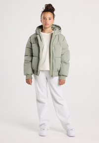 Joven con el cabello recogido en un moño, vistiendo una chaqueta acolchada color verde salvia, una camiseta beige, pantalones de chándal gris claro y zapatillas blancas, de pie frente a un fondo liso.