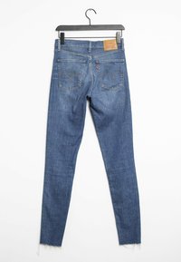 Jean skinny en denim bleu avec un ourlet effiloché, doté d'un patch en cuir marron et de deux poches arrière avec des coutures contrastantes.