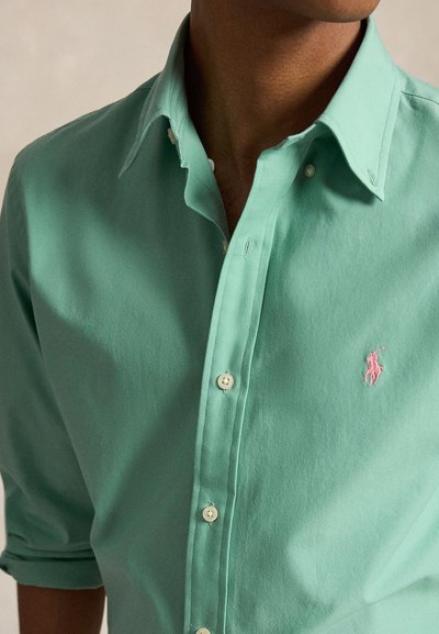 Mand iført en mintgrøn skjorte med knapper, opkørte ærmer og et lille lyserødt broderet logo på brystet.