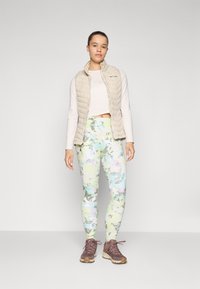 Lys beige pufvest over en hvid langærmet bluse, parret med pastelfarvede blomstrede leggings i grøn og blå. Brune vandrestøvler fuldender outfittet.