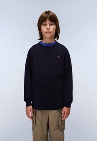 Felpa in cotone blu navy con collo rotondo, polsini a costine e un piccolo logo sul petto. Indossata sopra una camicia blu e pantaloni khaki.
