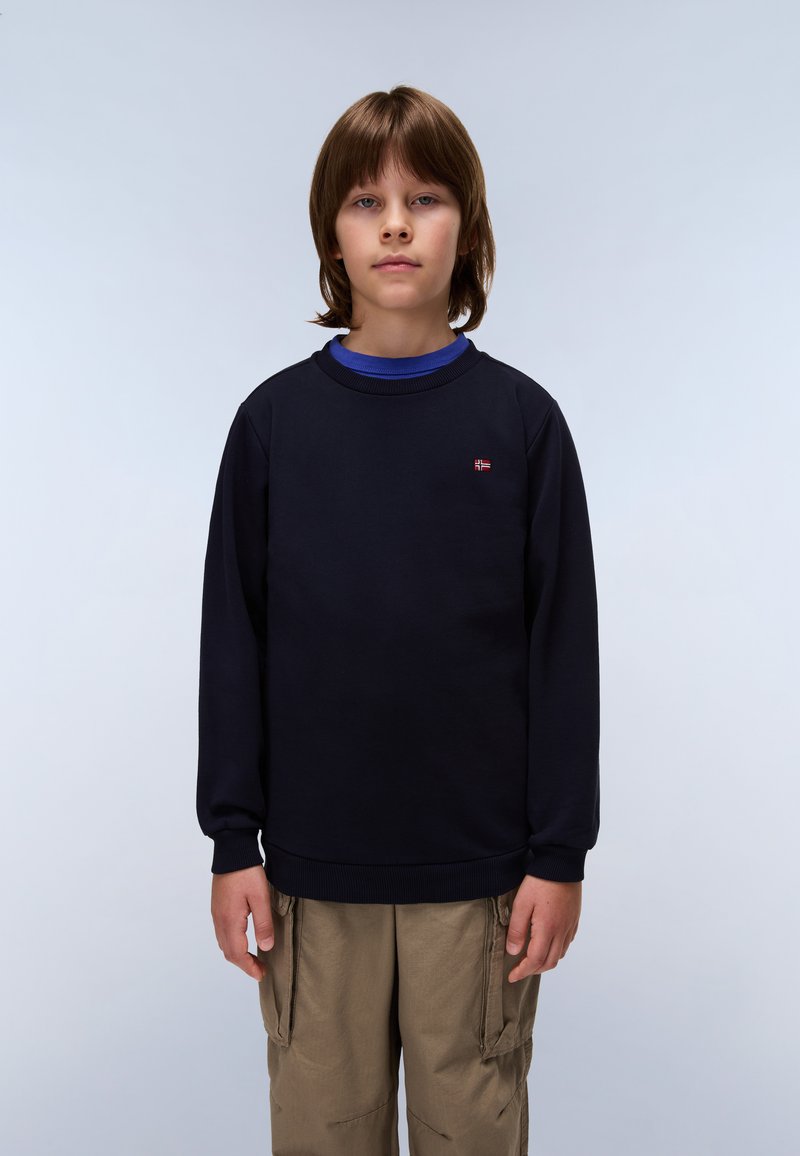 Felpa in cotone blu navy con collo rotondo, polsini a costine e un piccolo logo sul petto. Indossata sopra una camicia blu e pantaloni khaki.