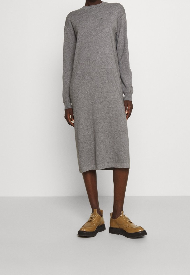 Personne portant une robe en maille grise à manches longues, longueur genoux, associée à des chaussures lacées couleur camel sur un fond blanc uni.