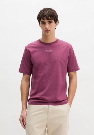 Młody mężczyzna o krótkich ciemnych włosach, ubrany w fioletową koszulkę Marc O'Polo i beżowe spodnie, stojący na tle jednolitego białego tła.