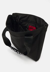 Sac fourre-tout noir avec de larges poignées, poche zippée intérieure et étiquette rouge visible affichant "HUGO" à l'intérieur du sac.