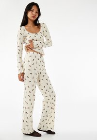 Vrouw in een crèmekleurig geribbeld shirt met lange mouwen en een broek met een klein bruin teddybeertje-print, staand met één hand op haar heup en donkere sokken.