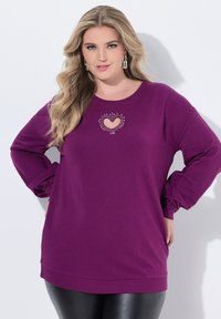 Donna con lunghi capelli biondi che indossa un maglione viola con taglio a cuore e dettagli di strass, mentre posa con le mani sui fianchi su uno sfondo bianco.