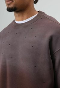 Bruine trui met een gladde textuur, ronde hals en verspreide zwarte versieringen over het oppervlak van de stof.