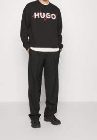 Sudadera negra con el logo "HUGO" en blanco y rojo, combinada con pantalones negros y zapatillas negras. Diseño simple y corte relajado.