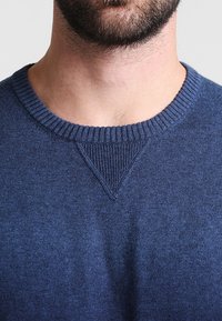 Marinblå tröja med rund halsringning, ribbade muddar och en texturerad tygkvalitet. Har en subtil V-stickningsdetalj vid halsen.