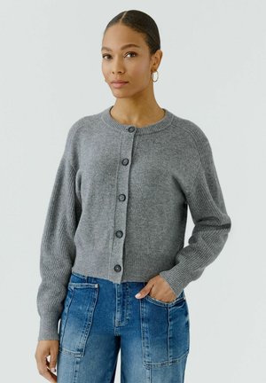 Frau mit Dutt trägt goldene Creolen, eine graue, geknöpfte Strickjacke und blaue Jeans mit großen Fronttaschen, steht vor einem schlichten Hintergrund.