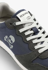 Baskets bleu marine et grises avec une tige en daim, des lacets blancs et un logo crâne et os croisés bien en vue. Accents texturés avec un motif en forme de ciseaux.