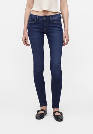Jean slim fit bleu foncé porté par une personne debout avec un top court blanc et des chaussures Mary Jane noires sur fond blanc.