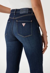Dark blue denim jeans with a fitted design, featuring a Guess logo patch on the back waistband and two back pockets with triangle accents.