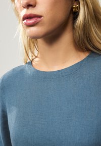 Top in lino azzurro chiaro con scollo arrotondato, tessuto strutturato e design minimalista. Dettaglio orecchino visibile.