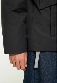 Svart jacka med en slät textur, med breda manschetter och ett grått dragband med fiskbensmönster. Bärs över blå denimbyxor.