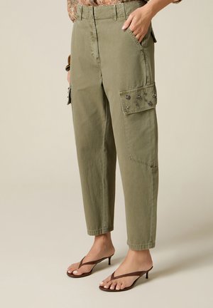 Pantalon cargo vert olive avec poches latérales embellies, porté avec des sandales marron à talons et tongs, main du modèle dans la poche, fond beige.