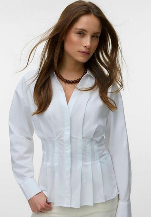 Vrouw met lang bruin haar draagt een getailleerde witte blouse met plooien en een donkere kralenketting tegen een effen lichte achtergrond.