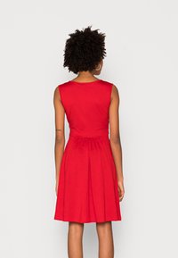 Robe rouge sans manches avec un corsage ajusté et une jupe évasée. Tissu lisse avec une finition naturelle, plis à la taille et encolure arrondie.