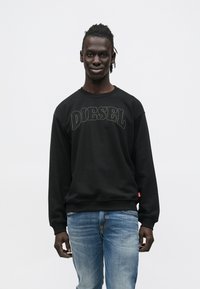 Μαύρο φούτερ με το λογότυπο "DIESEL" σε λευκή κεντητική, λαιμόκοψη crew, ριμπ μανσέτες, σε συνδυασμό με ξεθωριασμένο μπλε τζιν. Απλό φόντο.