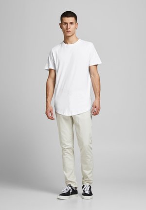 White cotton T-shirt with short sleeves, rounded hem, paired with light beige trousers and black sneakers featuring white laces.