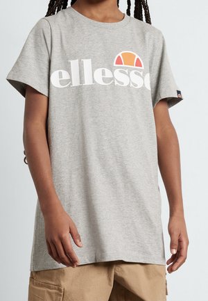 Osoba ubrana w jasnoszarą koszulkę Ellesse z pomarańczowo-czerwonym logo, połączoną z beżowymi szortami typu cargo, stoi na jednolitym tle.