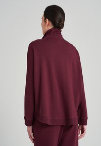 Bordó pulovr z měkkého materiálu, s vysokým límcem, kleslými rameny a žebrovanými manžetami. Jednoduchý design s volným střihem.