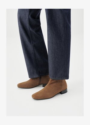 Bottines en daim marron avec bouts pointus et talons noirs bas, portées avec un jean large bleu foncé. La texture est lisse et douce.