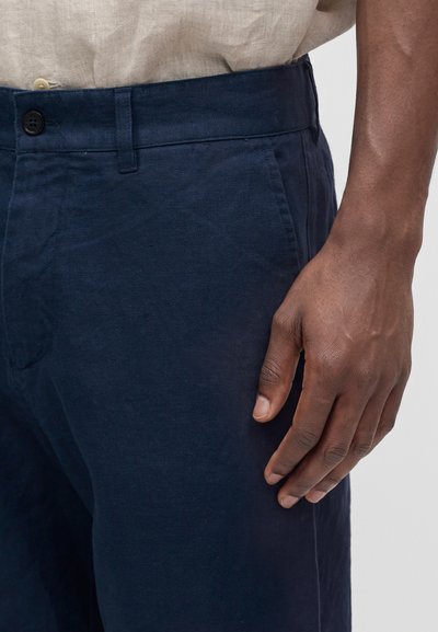 Homme portant un pantalon bleu marine et une chemise beige, main reposant à côté de la cuisse, montrant la texture du tissu et le détail du bouton.