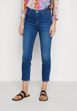 Kobieta w niebieskich, skróconych dżinsach, kolorowej bluzce z długim rękawem i czarnych sandałach na płaskim obcasie z odkrytymi palcami stoi na białej podłodze.