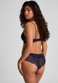 Vrouw met krullend haar, gekleed in marineblauwe kanten lingerie, met de rug naar de camera gekeerd en het hoofd naar de camera gedraaid tegen een effen grijze achtergrond.