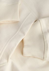 Μπεζ φανελάκι από ύφασμα με ριγωτούς μανσέτες και λεία υφή, με ορατές ραφές και ελαφρώς χαλαρή εφαρμογή.