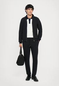 Melnā zip-up jaka virs baltā polo krekla ar tumši zilām malām, apvienota ar melniem biksēm ar auklēm un melnām kurpēm. Tur rokassomu.