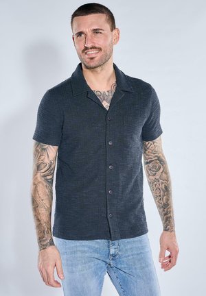 Mann mit kurzen Haaren und Tätowierungen auf beiden Armen und der Brust, trägt ein dunkelkurzes Hemd mit Knöpfen und hellblaue Jeans, leicht lächelnd.