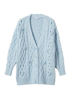 Vrouw met een zwarte rok, een zwart top met lange mouwen, een lichtblauwe grofgebreide cardigan en zwarte laarzen met hoge hakken tot aan de knie.