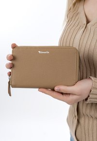 Käsivarret pitelevät vaaleanruskeaa, teksturoitua nahkaista vetoketjullista lompakkoa, jossa on "Tamaris"-logo. Kuvassa on myös beige, ribbineulottu neule yksiväristä valkoista taustaa vasten.