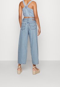 Světle modrý denimový crop top s křížovými ramínky v kombinaci s kalhotami s širokými nohavicemi. Obsahuje zadní kapsy a přírodní tkanou klínovou sandál.