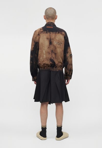 Άτομο με ξυρισμένο κεφάλι που φορά ένα καφέ και μαύρο φθαρμένο μπουφάν, μαύρη πλισέ φούστα, μαύρες κάλτσες και μπεζ παντόφλες, γυρισμένος με την πλάτη.