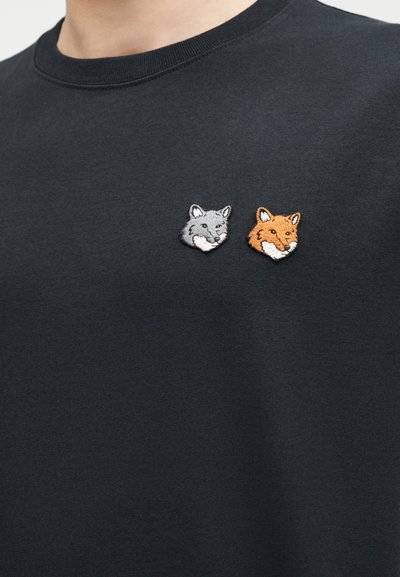 Μαύρο πουκάμισο με δύο κεντημένα κεφάλια αλεπούς, ένα γκρι και ένα πορτοκαλί, τοποθετημένα το ένα δίπλα στο άλλο στο πάνω μέρος του στήθους.