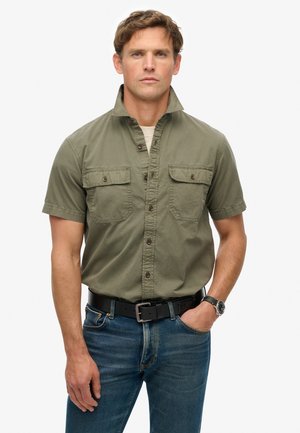 Olijfgroene overhemd met korte mouwen en knopen, met twee borstzakken, gedragen met blauwe jeans en een zwarte riem, met een casual kraagontwerp.