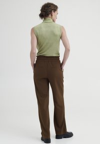 Hellgrüner ärmelloser Rollkragenpullover, kombiniert mit braunen, hochgeschnittenen Hosen mit Relaxed Fit und elastischem Bund. Schwarze Schuhe mit dicker Sohle.