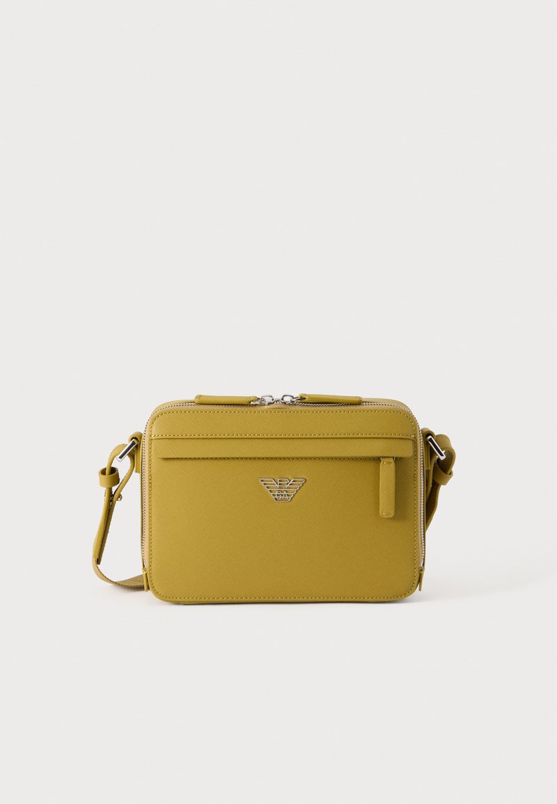 Small mustard yellow rectangular crossbody bag with front zipper pocket and silver logo emblem centered on front panel.