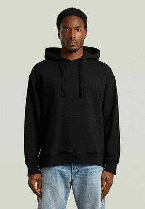 Uomo con felpa nera semplice e jeans azzurri, in piedi davanti a uno sfondo neutro, con espressione neutra e rivolto verso l'obiettivo.