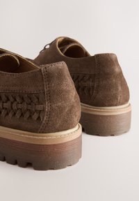 Chaussures en daim marron avec un motif tissé sur les côtés et des semelles épaisses et texturées. Le talon présente un design en couches de bois et de caoutchouc.