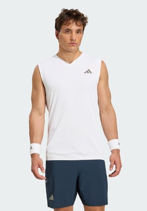 Mand iført hvid ærmeløs sportstrøje, marineblå shorts og hvide håndledsbånd, stående med fronten mod kameraet mod en ensfarvet baggrund.